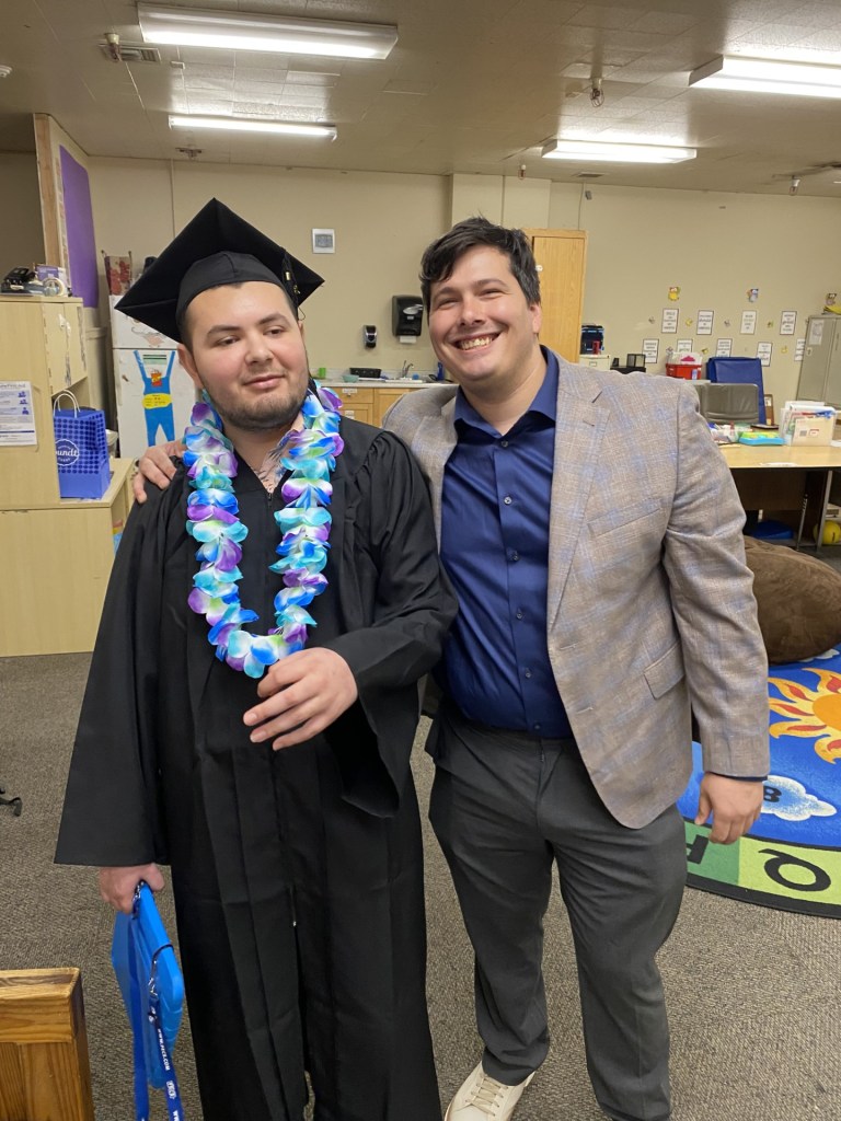 Lattice graduate seated with two supporters in front of a black and gold graduation backdrop.