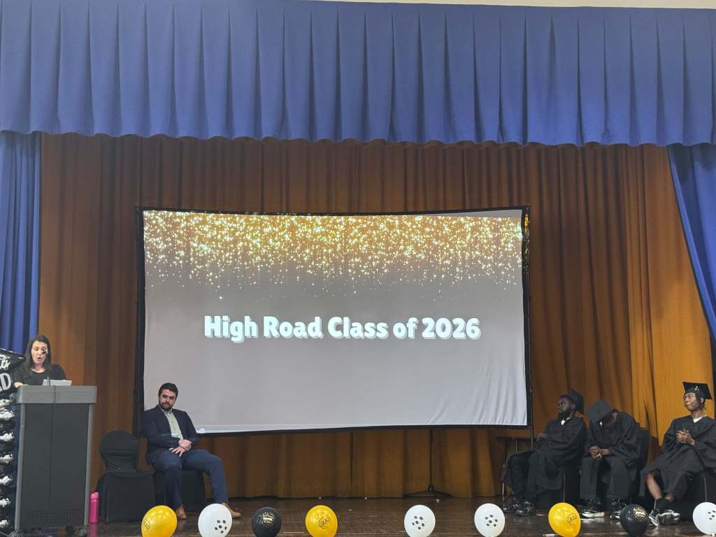 Ceremonia de graduación de la promoción de 2026 del programa High Road de Ferguson, con los graduados sentados en el escenario bajo una pantalla de proyección decorativa.