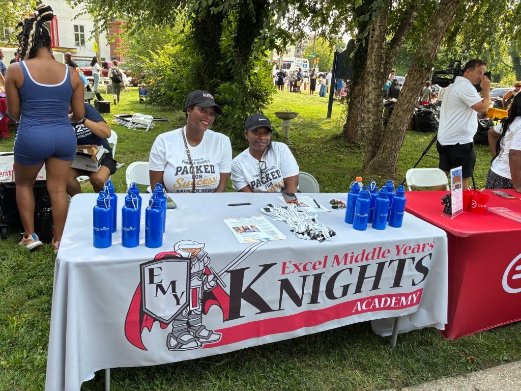 Excel Middle Years Academy staff at an information table during the Philadelphia back-to-school event, with school materials and branded water bottles on display.