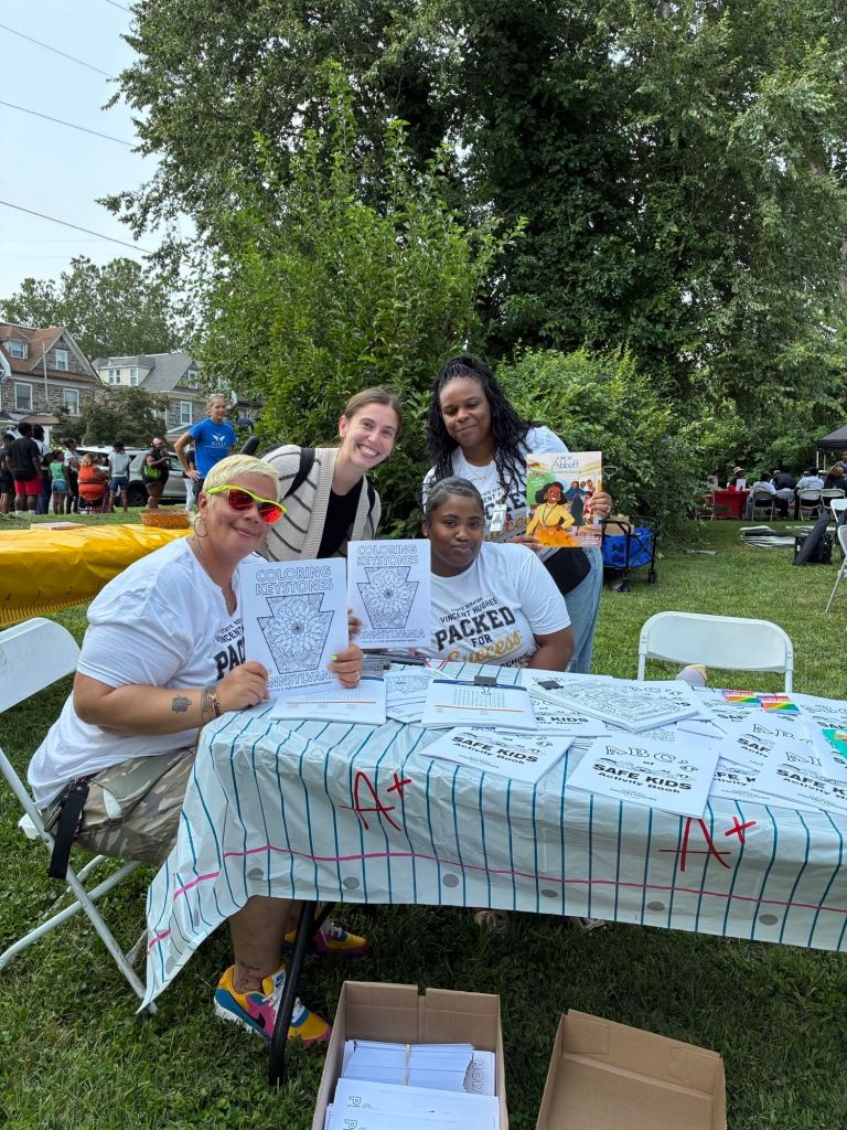 Community partners pose at an activity table with coloring pages and student resources during the Packed for Success back-to-school event in Philadelphia.
