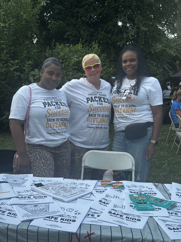 Packed for Success volunteers pose behind a table filled with coloring books, crayons and student activity materials.