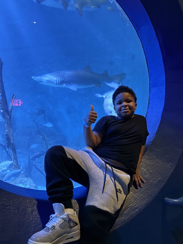 High Road School student gives a thumbs up while sitting beside a large aquarium window with sharks swimming behind him.