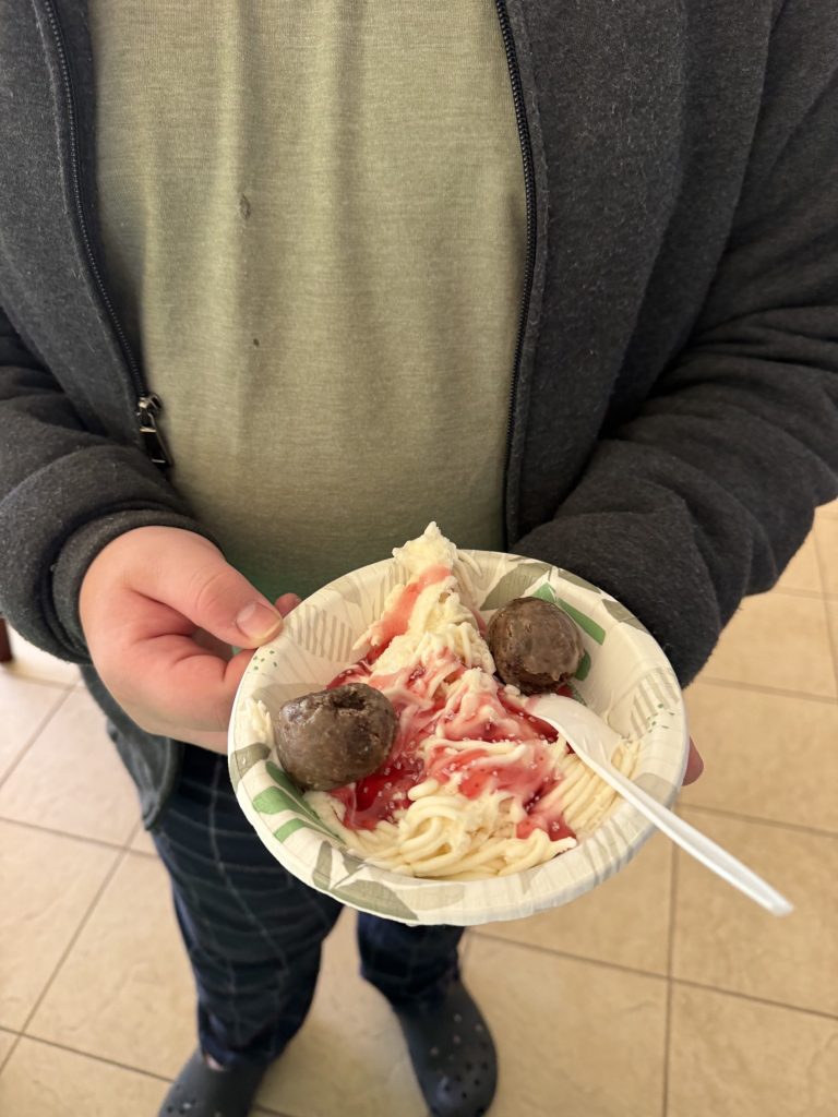A finished bowl of vanilla ice cream styled as spaghetti with strawberry sauce, white sprinkles and chocolate donut holes.