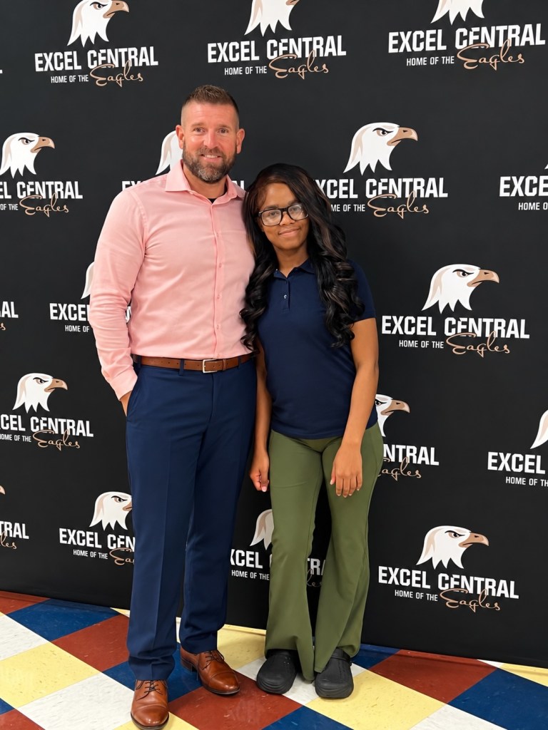 Arielle Johnson stands beside a man in a pink shirt in front of an Excel Central Eagles backdrop.