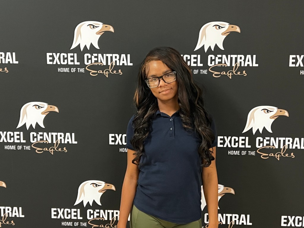 Excel Academy Central senior Arielle Johnson poses in front of an Excel Central Eagles backdrop, wearing a navy polo shirt and glasses.