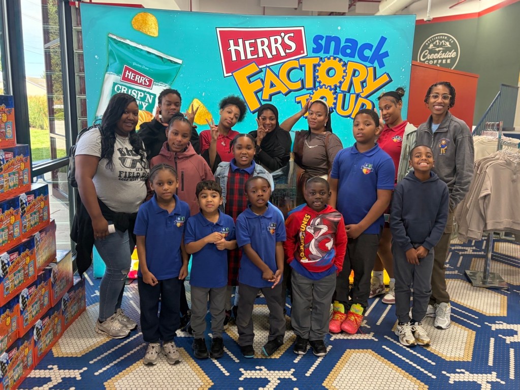 High Road School at Chester Community Charter School students pose outside the Herr’s factory beside a large red wall displaying the company logo.