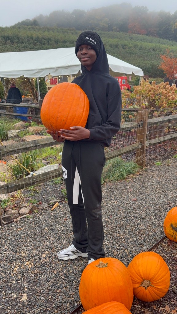 Un alumno de la High Road School del condado de Fairfield, vestido con una sudadera negra con capucha y un gorro de punto, sostiene una gran calabaza naranja en la granja Silverman's Farm.