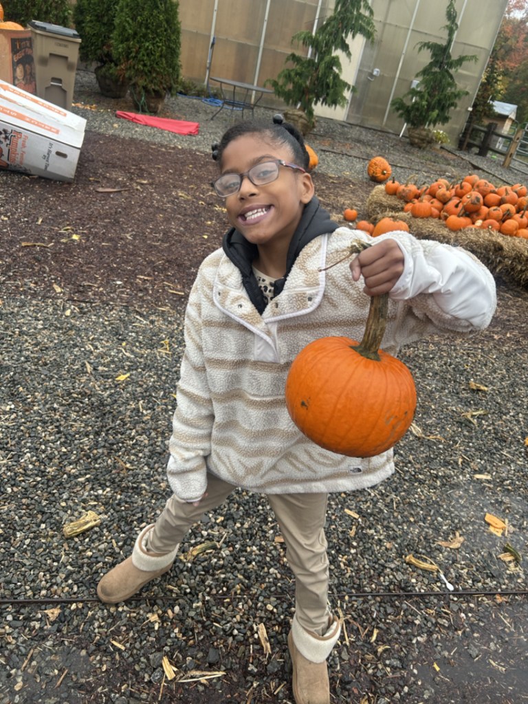 Un alumno de la escuela Smiling High Road del condado de Fairfield, que lleva gafas y una chaqueta de forro polar de color crema, sostiene con orgullo una calabaza.