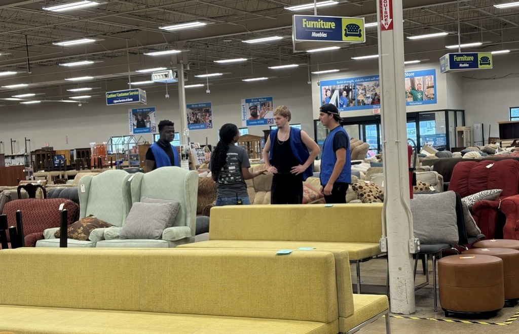 Students in blue volunteer vests speak with another person among sofas and chairs in the Habitat for Humanity ReStore furniture section.