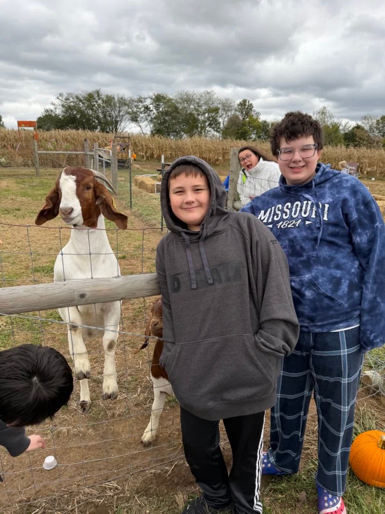 Dos alumnos de la escuela High Road de Wright City sonríen para una foto junto a una cabra en el huerto de calabazas.