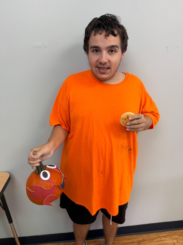 High Road School of Hoffman Estates Fall Fest participant wearing an orange shirt displays a decorated pumpkin while holding a cookie.