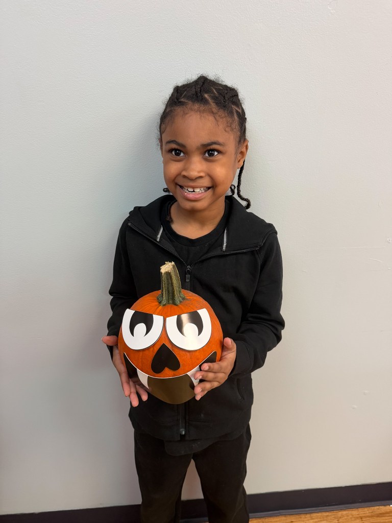 Smiling High Road School of Hoffman Estates student wearing black holds a pumpkin decorated with large cartoon eyes and a monster face.