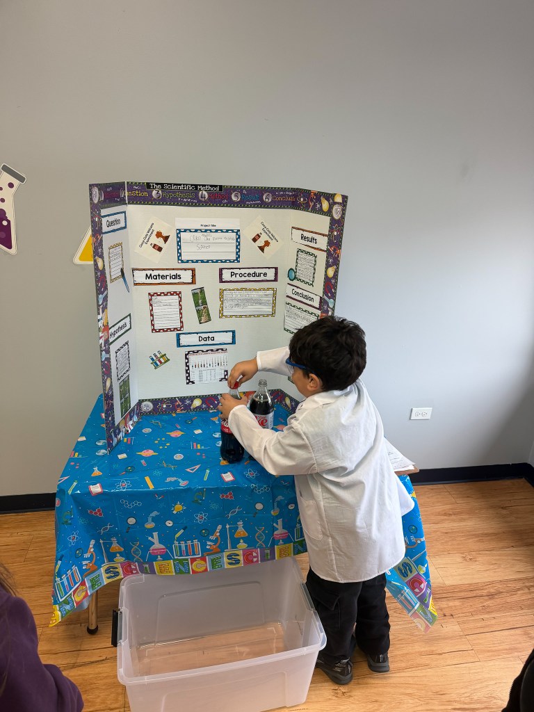 Un alumno de la High Road School de Hoffman Estates, vestido con una bata de laboratorio, prepara un experimento con Coca-Cola Light y Mentos junto a un panel de exposición de la feria de ciencias.