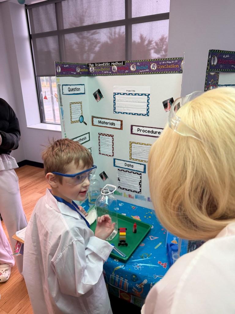 Un alumno de la escuela Young High Road de Hoffman Estates, que lleva gafas de seguridad, comenta un experimento científico con un adulto junto a un panel expositivo.