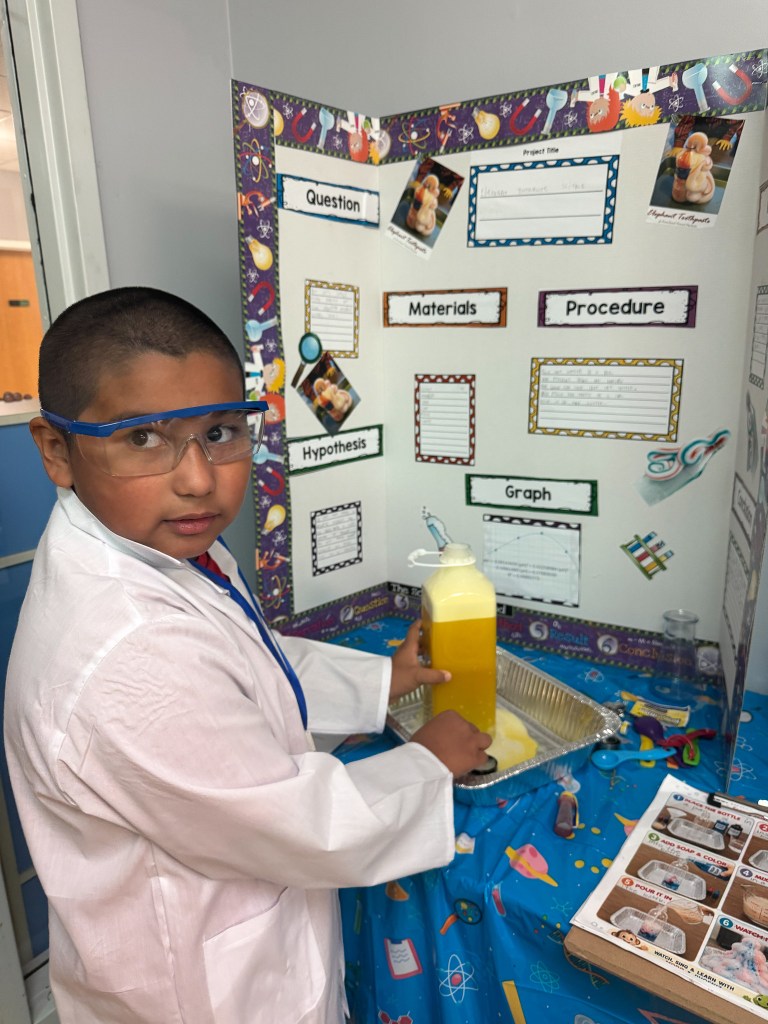Un alumno de la High Road School de Hoffman Estates, vestido con bata de laboratorio y gafas de seguridad, sostiene una botella llena de un líquido amarillo junto a un proyecto de la feria de ciencias.