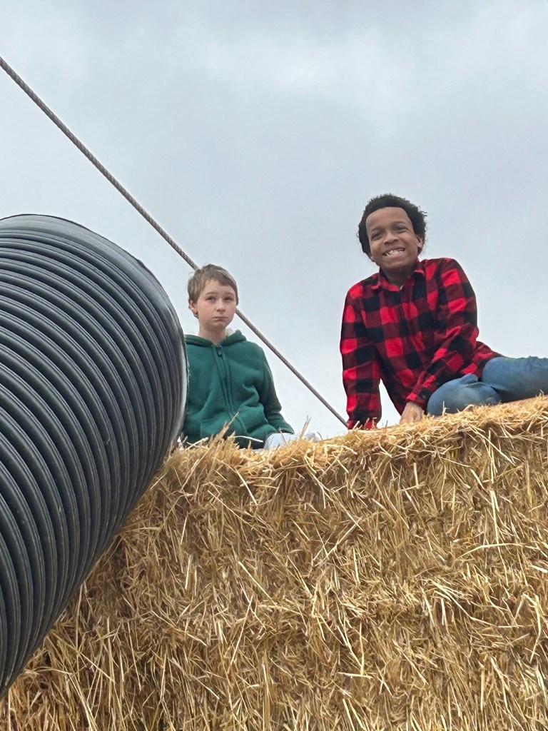 Dos estudiantes están sentados sobre fardos de heno junto a un gran tubo negro durante una visita a la granja Lincoln Ridge.