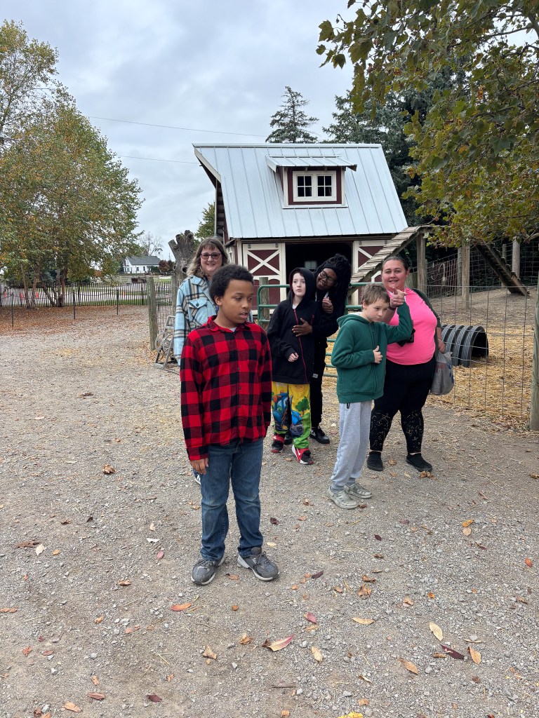 Los alumnos y los adultos se reúnen al aire libre cerca de un pequeño granero durante una excursión de otoño.