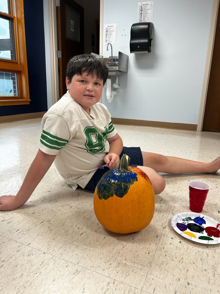 Un alumno está sentado junto a una calabaza mientras participa en la actividad de pintura de calabazas en la escuela High Road del condado de Cole.